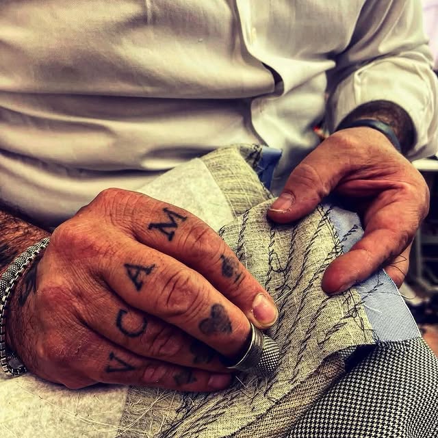 Le mani tatuate di Marco, ditale al dito, che imbastiscono a mano il revers di una giacca