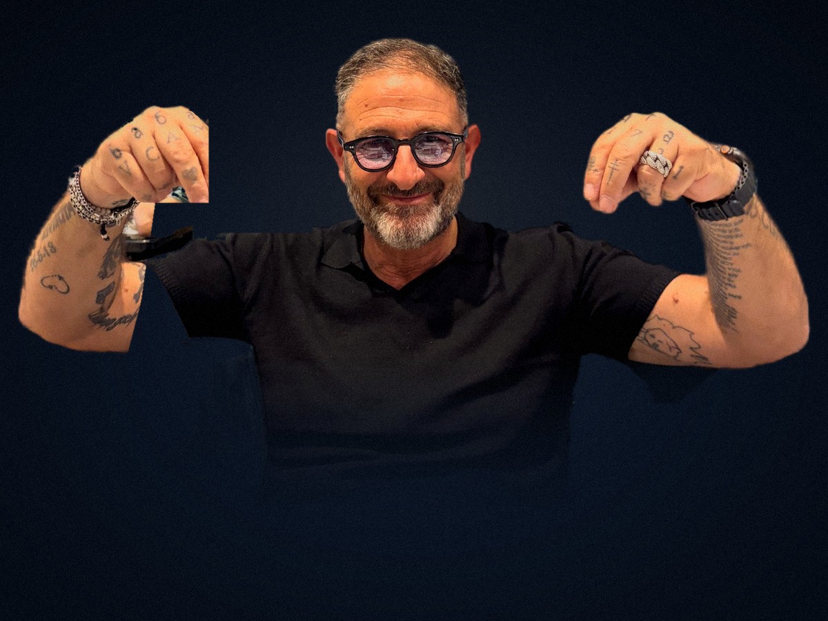 Marco Parroni nel suo atelier: occhiali, barba sale e pepe, polo nera e pugni tatuati alzati, con un sorriso